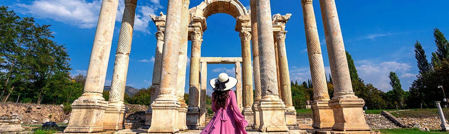 A person in a long purple dress and white hat standing amidst ancient, tall, white columns under a clear blue sky.