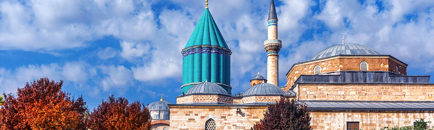 A historic mosque with a prominent turquoise dome and a tall minaret, surrounded by autumn trees under a blue sky with fluffy clouds.