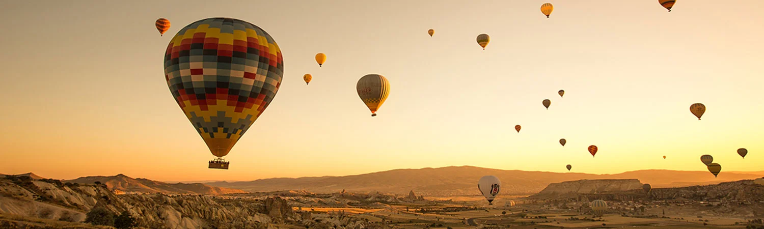 A scenic view of multiple colorful hot air balloons floating in the sky at sunset, with a picturesque landscape of mountains and valleys below. Keywords: Cappadocia, Turkey, balloon ride, fairy chimneys, UNESCO World Heritage Site.
