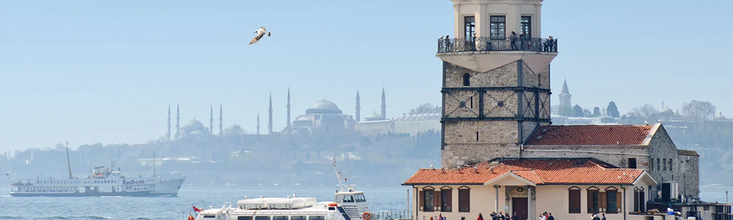 A panoramic view of the Bosphorus Strait featuring the Maiden’s Tower in the foreground, boats on the water, and a distant view of Istanbul’s skyline with mosques and minarets under a clear blue sky.
