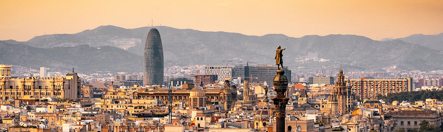 A sunset view of Barcelona’s cityscape with the Torre Glòries, a modern oval-shaped building, and traditional buildings in the foreground.