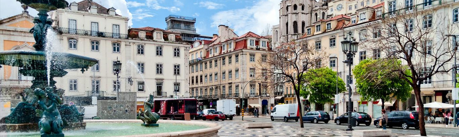 A busy street scene in Lisbon featuring a large fountain, classic buildings, and vehicles parked along the sidewalk.