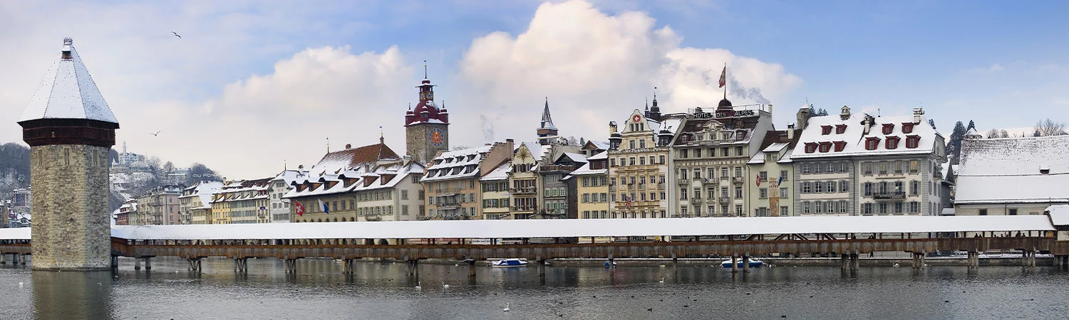 “Panoramic view of a snowy European cityscape with historical architecture, a stone tower, and a wooden bridge over a calm river.”