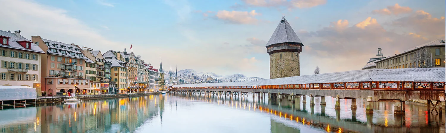 A picturesque winter scene of the Chapel Bridge and Water Tower in Lucerne, Switzerland, with snow-covered rooftops and serene reflections on the calm water.