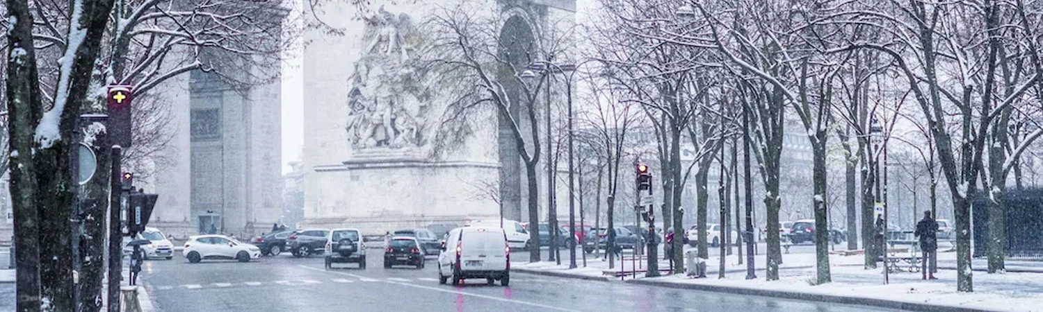 A snowy city street with cars, bare trees, and a large stone monument in the background