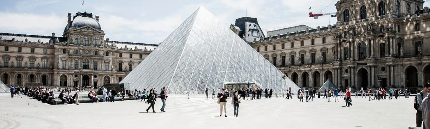 Tourists visiting the iconic Louvre Museum courtyard featuring the famous glass pyramid and historic architecture in Paris, France.