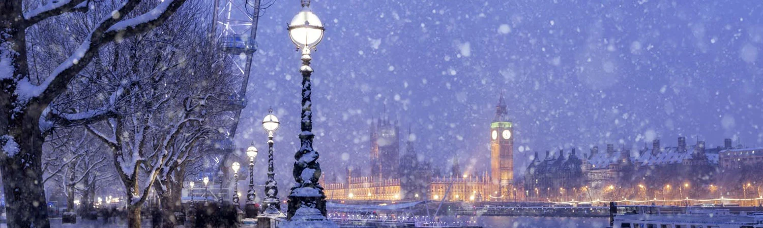 The image depicts a snowy evening in London. The scene is illuminated by street lamps casting a warm glow on the falling snowflakes.