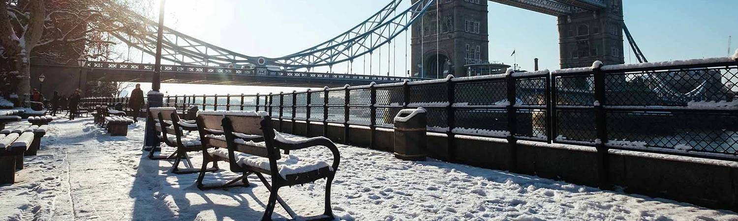 The image shows a serene, snowy scene near the iconic Tower Bridge in London. Several benches covered with snow are aligned along a walkway, with footprints visible, indicating recent activity.