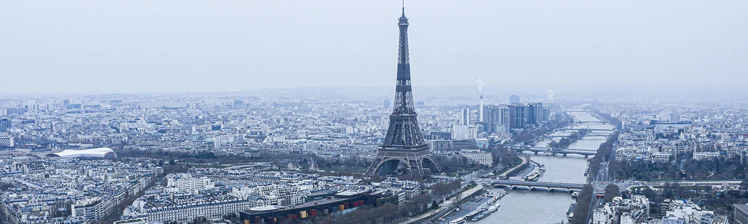 The image provides a panoramic view of Paris on a cloudy day. The Eiffel Tower stands tall amidst a vast cityscape, serving as the central focus of the image.