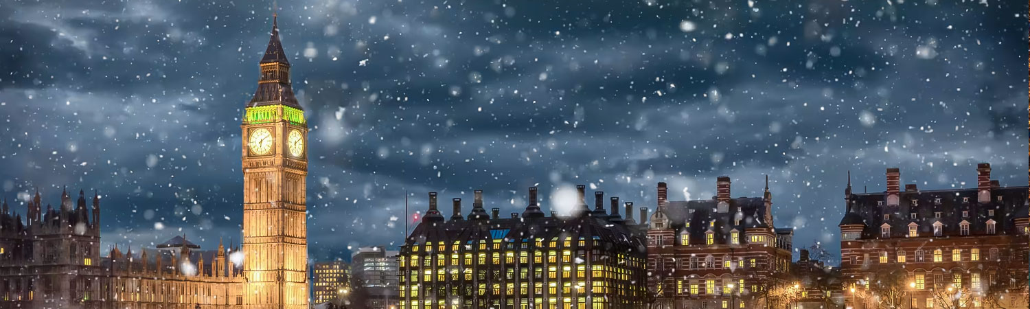 Big Ben and surrounding buildings in London on a snowy night. The clock face of Big Ben is illuminated with a warm light, while the surrounding buildings are also lit up, showcasing their architectural details.