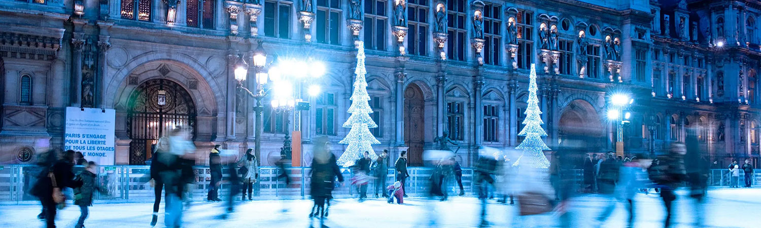 People ice skating at night in front of a beautifully illuminated historic building, with sparkling Christmas tree lights adding to the festive atmosphere. 