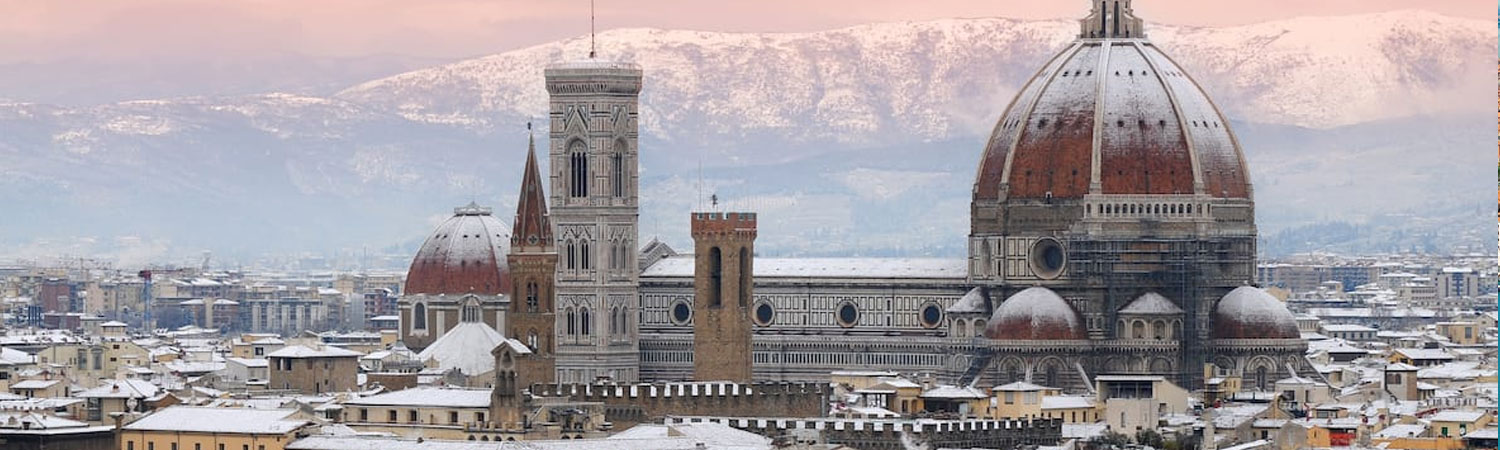 Panoramic view of Florence Cathedral with its iconic dome and bell tower, amidst a cityscape of snow-covered roofs, backed by mountains.