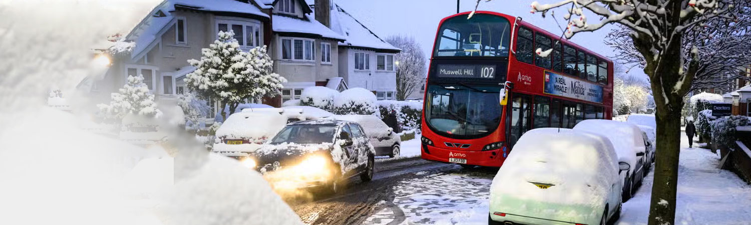 A winter day in London, where a red bus travels on a snow-covered road, passing by cars and houses.