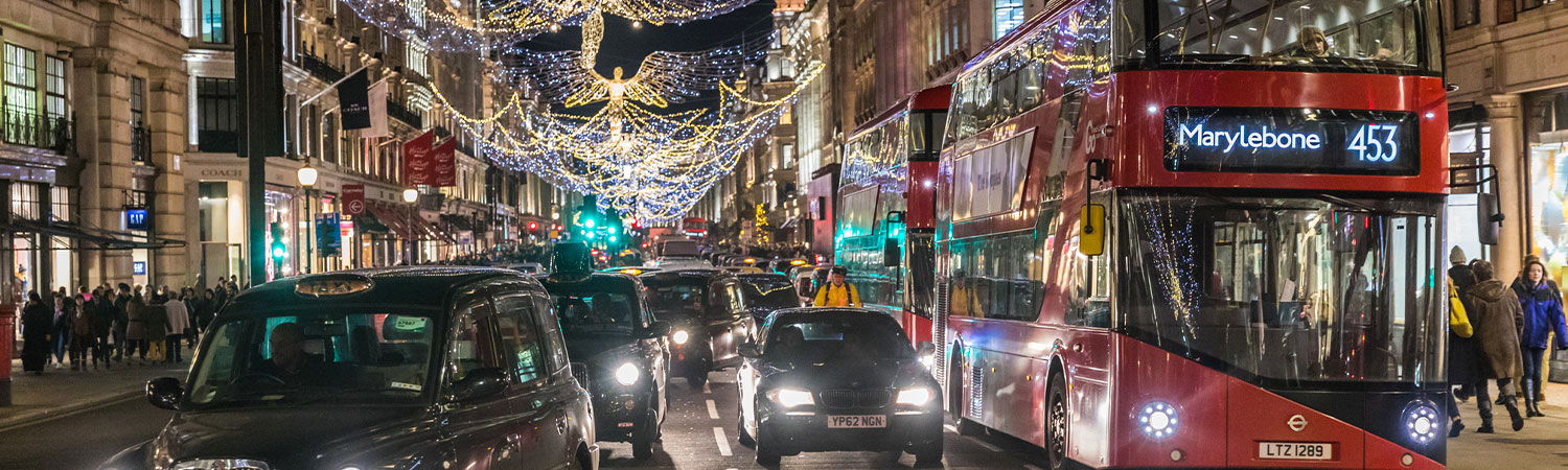 A bustling London street at night, illuminated by intricate holiday lights, with a red double-decker bus marked ‘Marylebone 453’, black cabs, and pedestrians.