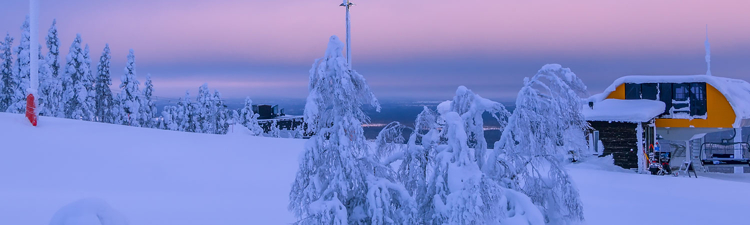 Snow-covered landscape at dusk with frosty trees, a ski lift, and a cabin against a pastel sky.
