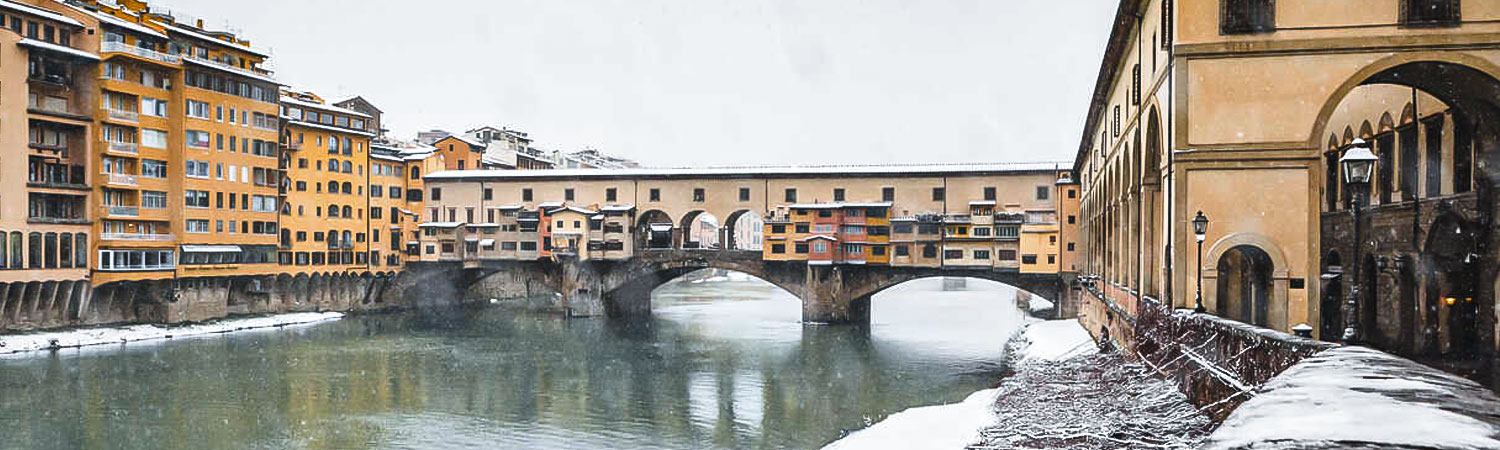 Snowy scene of the Ponte Vecchio, an iconic medieval stone bridge with shops built along it, spanning over the Arno River in Florence, Italy.