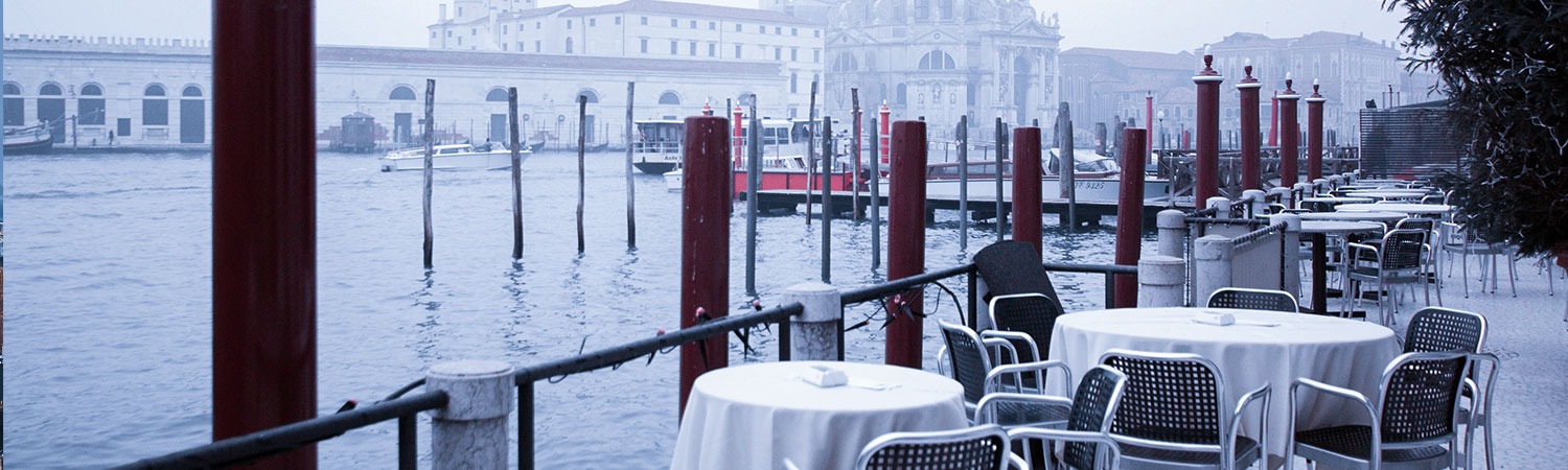 Serene outdoor dining area with white tablecloths and metal chairs overlooking a misty harbor, with red mooring poles and historic architecture in the background.