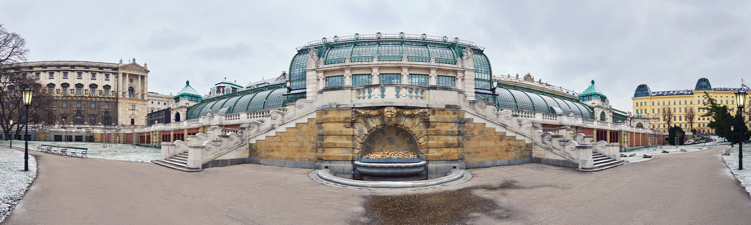 A historic building with a curved facade, a fountain, and green glass structures on the roof, in a classical cityscape under an overcast sky.