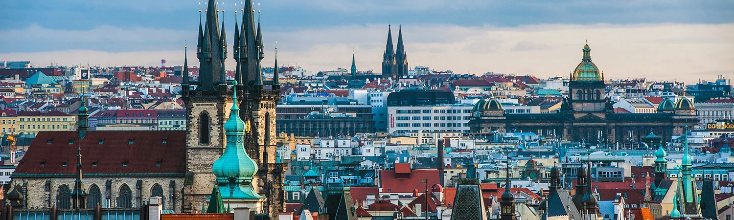 A panoramic view of Prague, showcasing the historic churches with towering spires and the sea of red rooftops under a cloudy sky.