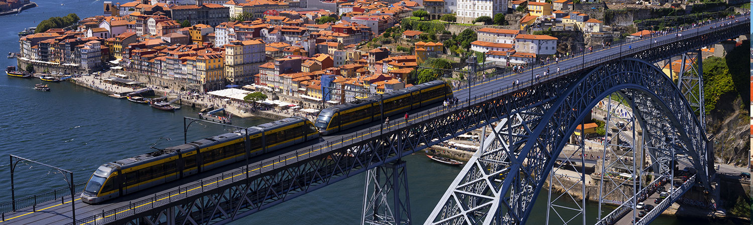 A yellow train crossing the Dom Luis I Bridge in Porto, Portugal, with the Ribeira District in the background on a clear day.