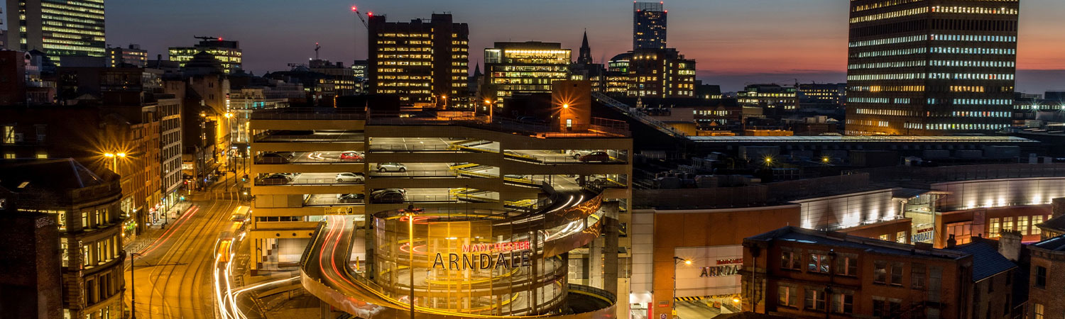 An evening view of Manchester, showcasing the vibrant city lights, the Arndale Centre, and modern architecture under a dusk sky.
