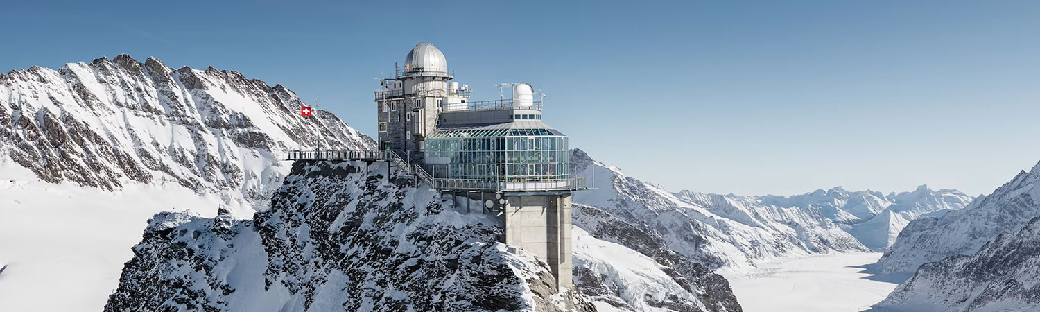 The image captures a breathtaking view of a modern observatory perched on the rugged cliffs of the Swiss Alps, overlooking a pristine snow-covered valley.