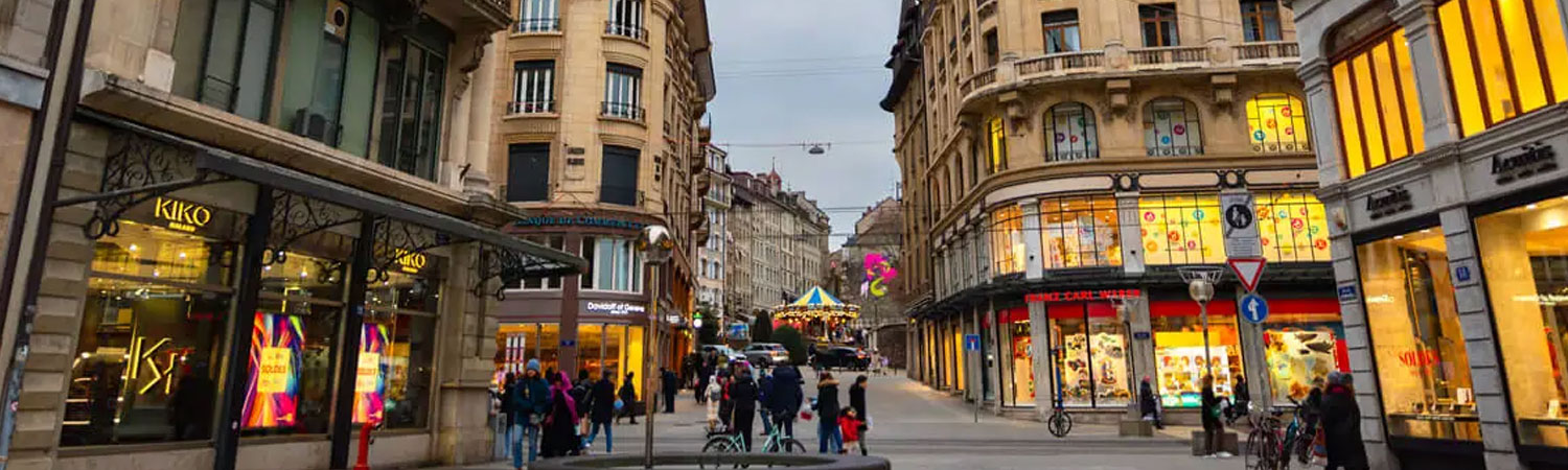 The image captures a bustling street scene in the evening at a popular shopping district in Geneva.