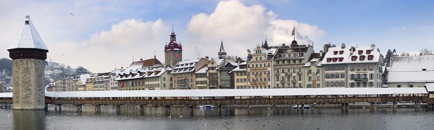The image captures a panoramic view of a snowy landscape featuring the iconic Chapel Bridge in Lucerne, Switzerland. 