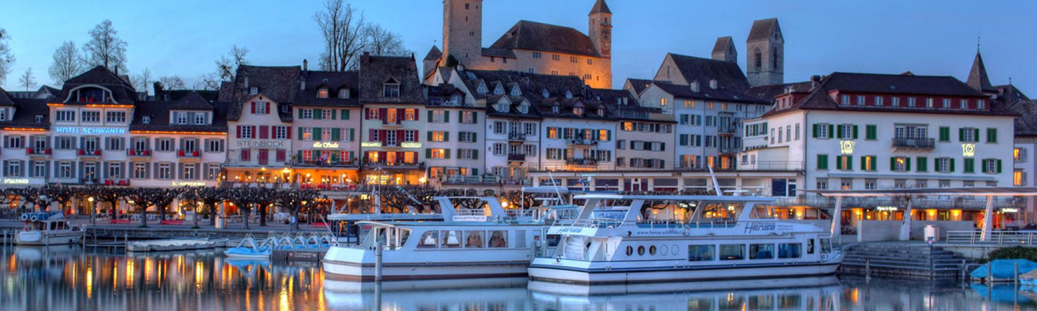 A scenic evening view of a European waterfront town with illuminated buildings, a calm harbor, and moored boats reflecting on the water.