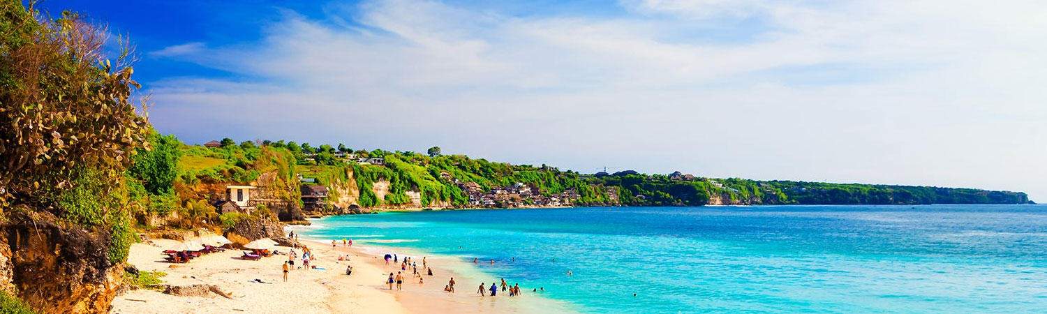 Bali beach with turquoise water, sandy shore, cliffside homes and people relaxing.