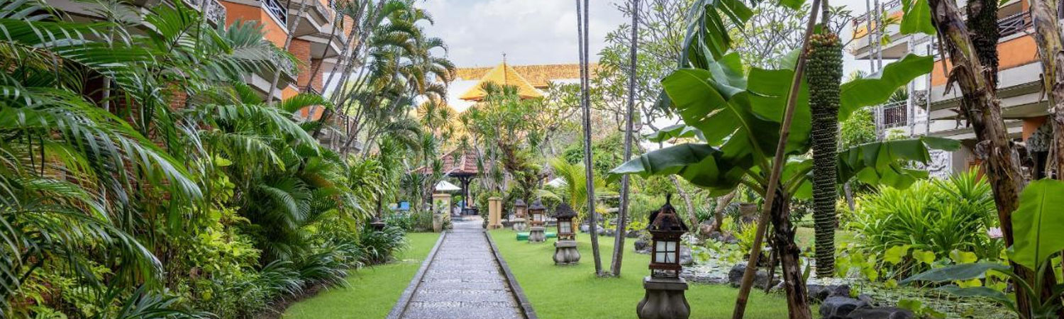 Garden pathway with tropical plants, lanterns and golden-roofed building in a resort setting.
