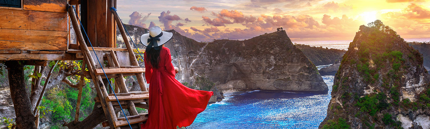 A person in a flowing red dress and wide-brimmed hat admires the breathtaking view of the ocean and rocky cliffs during sunset from a wooden treehouse in Nusa Penida, Bali.