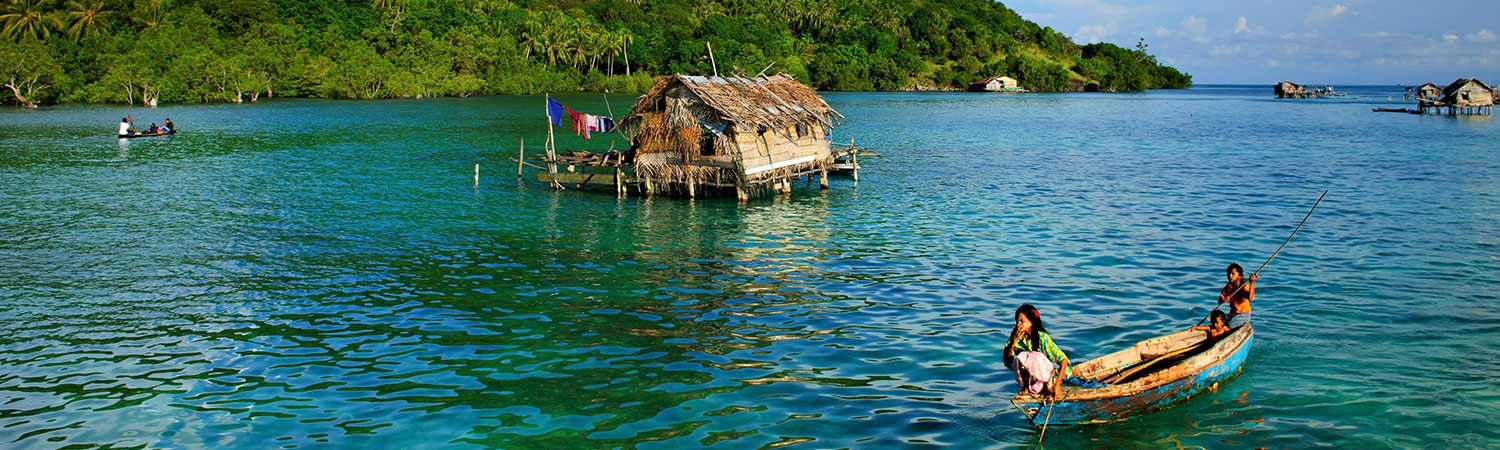 Scenic view of a tranquil tropical lagoon at an undisclosed location, featuring a traditional wooden boat with people, and overwater huts surrounded by lush greenery.