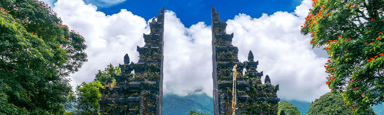 Panoramic view of the iconic Balinese Handara Gate, surrounded by lush greenery and vibrant flowers, with misty mountains and clouds in the background.