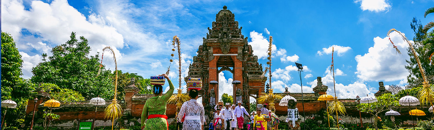 Vibrant ceremony at a traditional Balinese temple in Indonesia, with participants in colorful attire and decorative umbrellas, set against a backdrop of a clear sky.