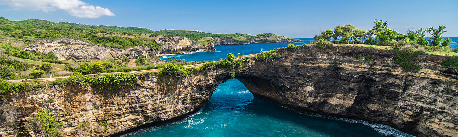 Breathtaking view of the natural stone bridge over turquoise waters at Broken Beach, Nusa Penida, Bali, surrounded by lush greenery and rugged cliffs under a clear blue sky.”
