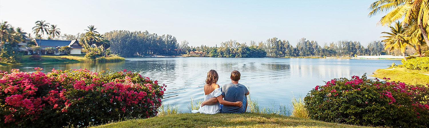 Two people enjoying a peaceful moment by a serene lake, surrounded by lush greenery and blooming pink flowers