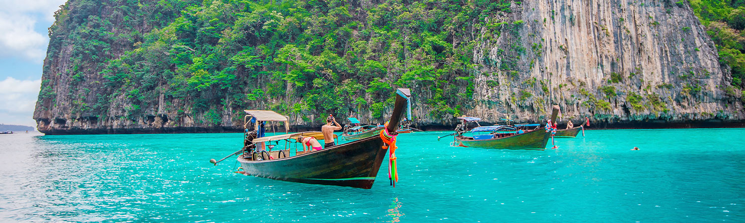 Traditional longtail boats floating on the crystal clear waters of a tropical beach in Thailand, near a lush, green limestone cliff under a partly cloudy blue sky.