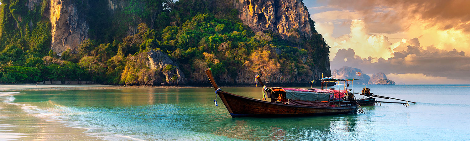 Traditional wooden boat anchored on a serene tropical beach in Thailand, with lush green cliffs and a golden sunset in the background.