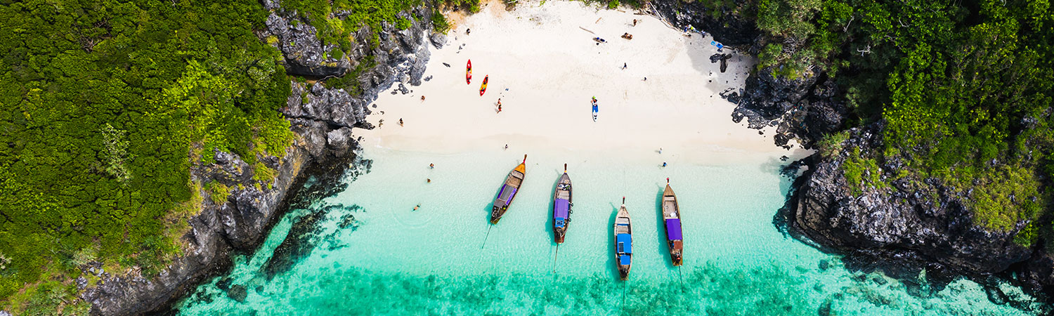 Aerial view of a pristine beach in Phi Phi Island, Krabi, Thailand, with turquoise waters, colorful boats anchored near the shore, surrounded by lush green cliffs.