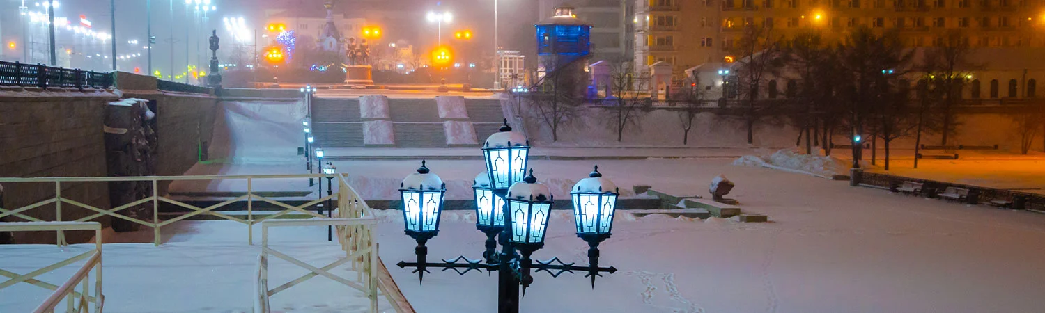A snowy evening in Azerbaijan with illuminated street lamps, a snow-covered park, and city lights in the background.