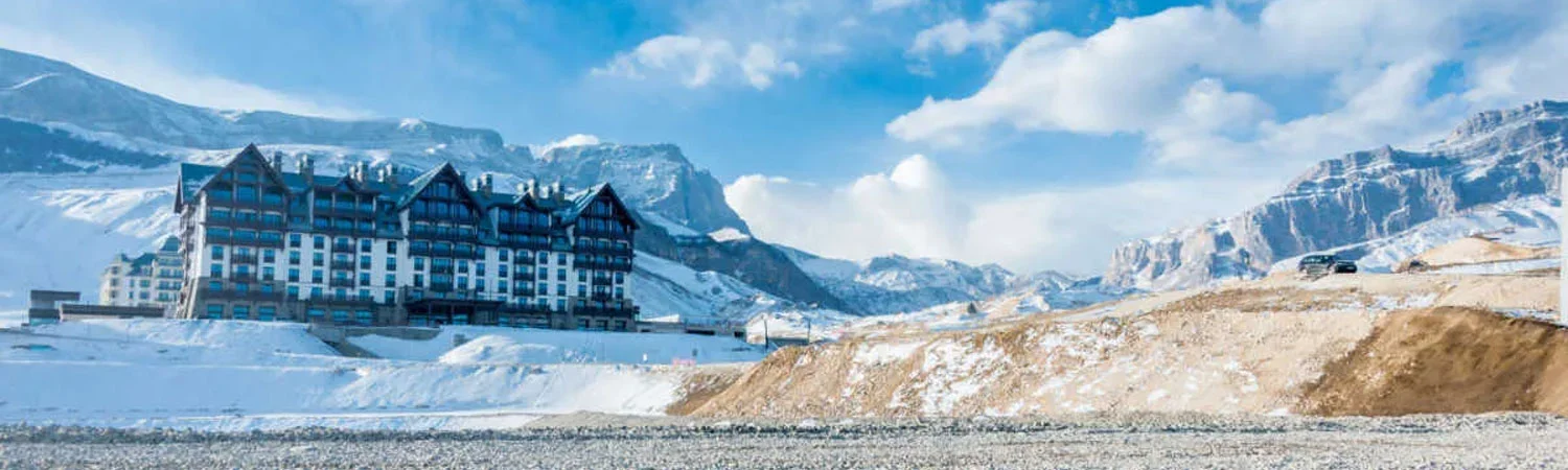 A large hotel with multiple balconies sits at the base of a snowy mountain range, under a partly cloudy sky. 