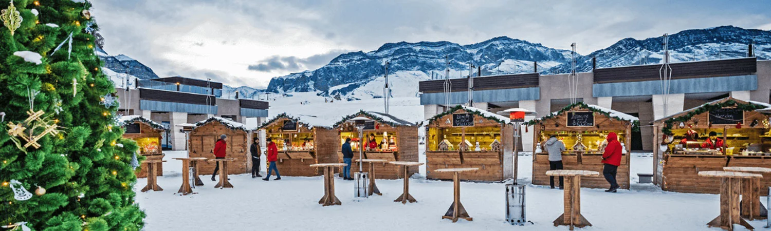 A snowy outdoor market in Azerbaijan with wooden stalls, people shopping, a decorated Christmas tree, and mountainous backdrop.