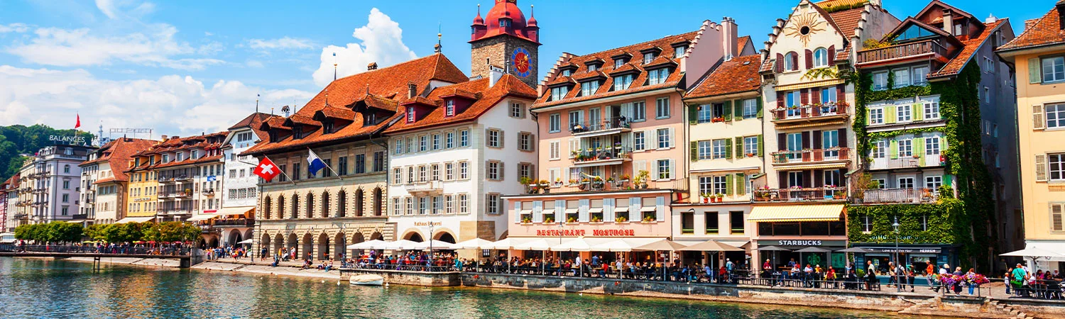 Vibrant waterfront scene in Lucerne, Switzerland, featuring colorful traditional Swiss architecture, bustling cafes including Starbucks, and people enjoying a sunny day along the promenade with green hills in the background