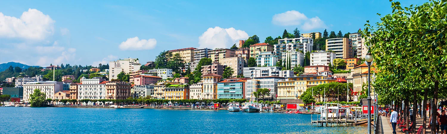 Panoramic cityscape with colorful buildings along a calm blue waterfront, docked boats, and a lively tree-lined promenade with people enjoying a sunny day, set against a backdrop of hills.