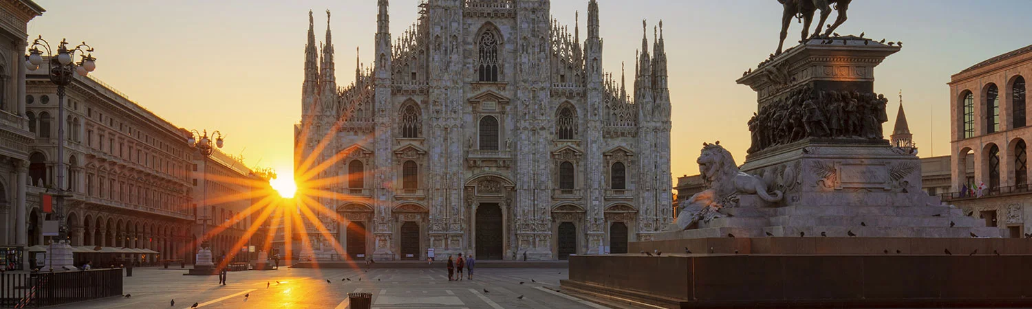 Sunrise at Milan Cathedral in Piazza del Duomo, showcasing the intricate Gothic architecture of the cathedral and surrounding buildings, with an equestrian statue and people in the distance under a clear sky.