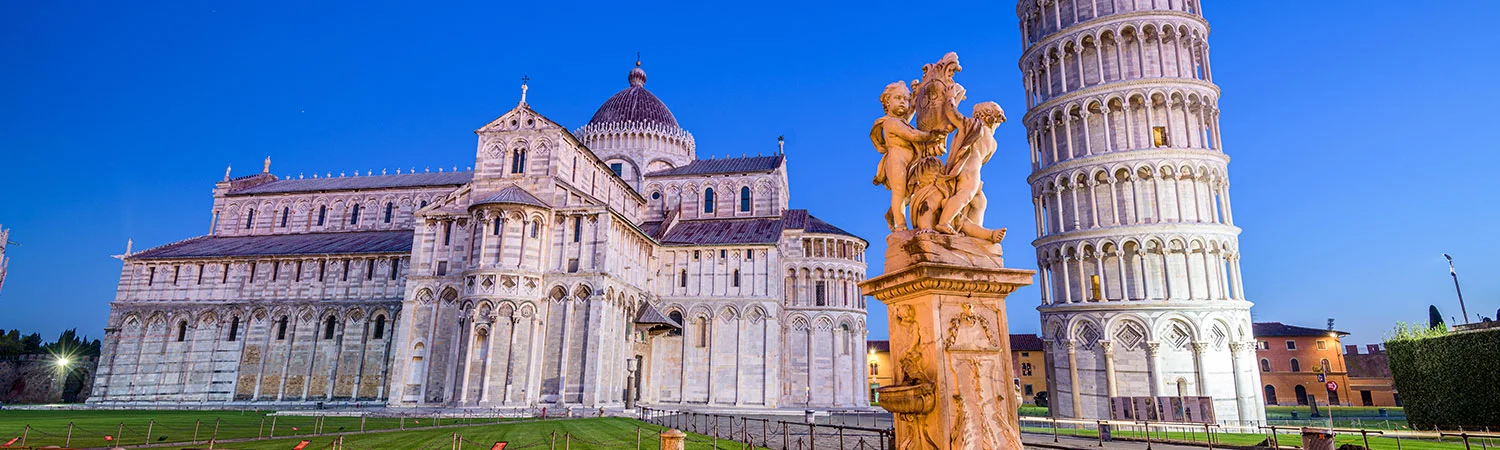 Panoramic view of the Pisa Cathedral and the Leaning Tower of Pisa illuminated at dusk, with a detailed stone statue in the foreground under a transitioning sky.