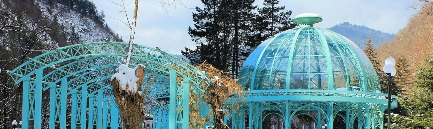 A scenic view of a turquoise iron gazebo covered in snow, located in a serene winter park with snow-capped trees and mountains in the background.