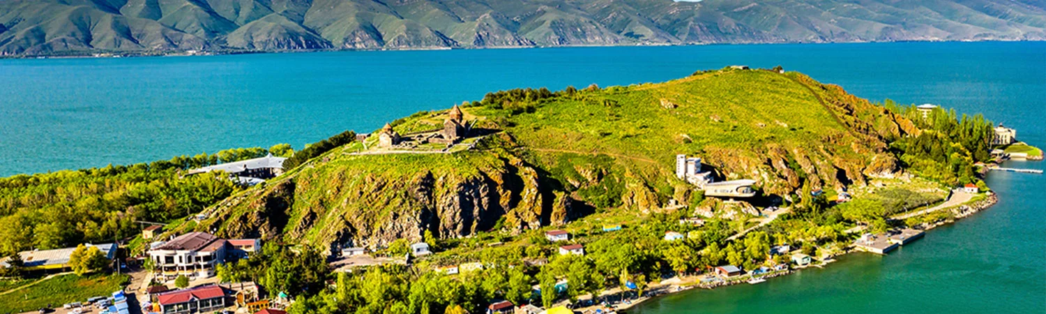 A panoramic view of a lush green hillside with buildings and ruins, bordered by a serene blue lake and distant mountains.