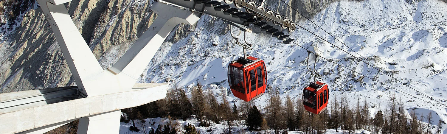 The image shows two red cable cars suspended in the air, moving along cables. A large concrete structure supports the cables from which the cable cars are hanging. 